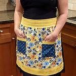 a woman standing in a kitchen wearing a yellow and blue flowered apron with pockets