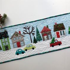 a quilted wall hanging with houses and cars on it's side, along with pine branches