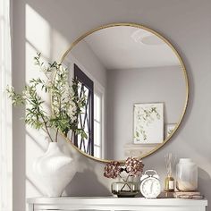 a white dresser with a round mirror on it's face and flowers in the vase