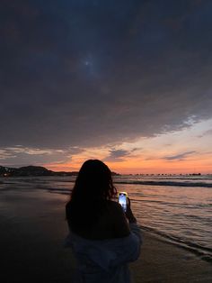 a woman standing on the beach taking a photo with her cell phone at sunset or dawn