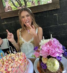 two women sitting at a table with cake and flowers in front of them, making the peace sign