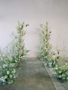 an arrangement of white flowers and greenery in front of a wall with concrete floor