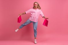 a woman in pink shirt and jeans holding shopping bags with both hands while standing on pink background