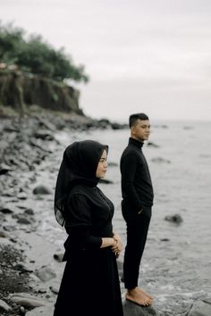 a man and woman standing next to each other on a beach near the ocean with rocks