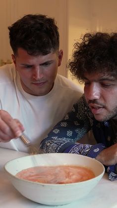 two men sitting at a table with a bowl of soup and a spoon in front of them