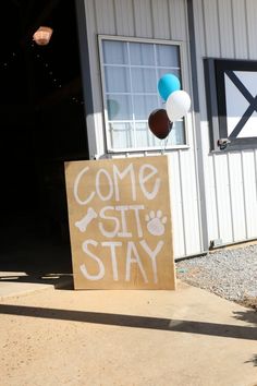 a sign that says come sit stay with balloons in front of a building and door