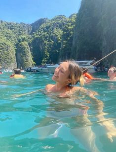 people swimming in the water near mountains and boats on the water, with one woman wearing an orange life jacket