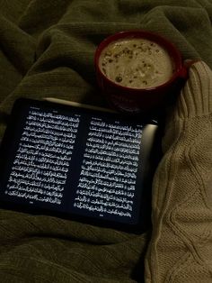 an electronic device sitting on top of a bed next to a cup of coffee and a book