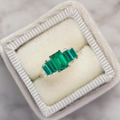 an emerald colored ring sits in a white box on a marble surface with silver trim around the edges