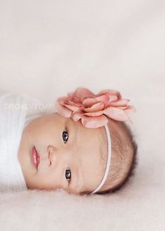 a baby laying down wearing a white shirt and flower in it's hair, looking at the camera