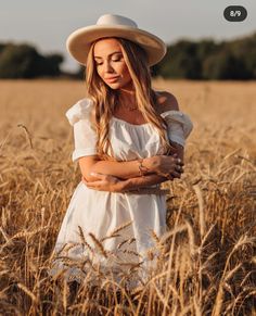 a woman in a white dress and hat standing in a wheat field with her arms crossed