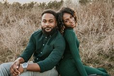 a man and woman sitting on the ground in front of tall grass