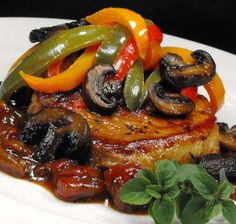 a white plate topped with meat covered in mushrooms and peppers on top of sauce next to green leaves