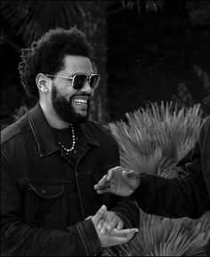 two men standing next to each other in front of trees and bushes, one holding his hand out