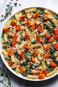 a white bowl filled with pasta and vegetables on top of a table next to herbs