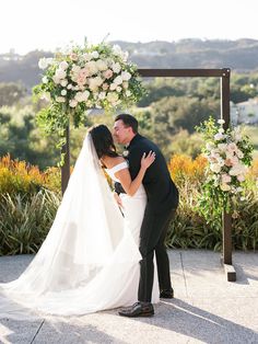 a bride and groom kissing under an arch decorated with white flowers at their wedding ceremony