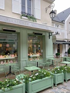 the outside of a store with green tables and chairs in front of it, surrounded by potted plants