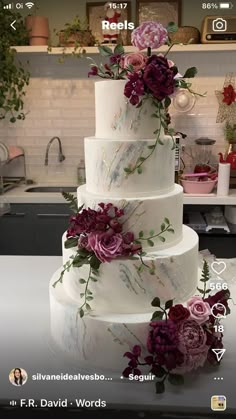 a three tiered wedding cake with purple flowers on the top and bottom, sitting on a kitchen counter