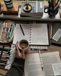 a person holding a coffee cup in their hand while sitting at a desk with books, pens and pencils