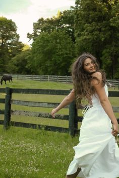 a woman in a white dress is walking through the grass near a fence and horses