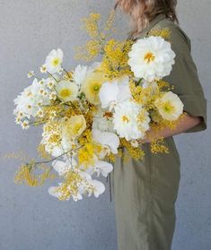 a woman holding a bouquet of white and yellow flowers in front of a gray wall