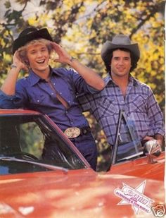 two men standing next to each other in front of a red car wearing cowboy hats