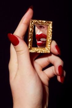 a woman with red nails holding up a golden square shaped object in front of her face