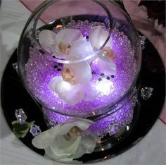 purple and white flowers are in a glass bowl on a black platter with crystals