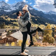 a woman holding a child in her arms while standing on a deck with mountains in the background