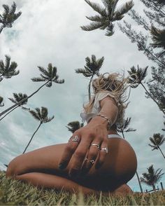 a woman sitting in the grass with her hands on her knees and palm trees behind her