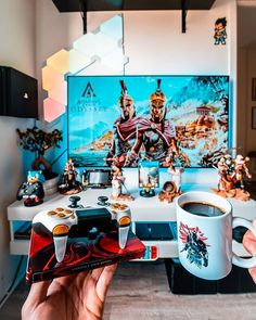 a person holding up a game controller in front of a desk with coffee and figurines on it