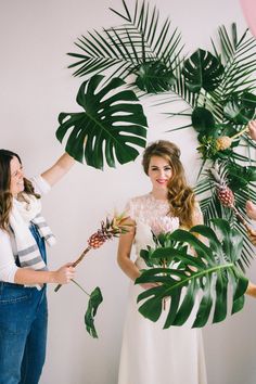 two women standing next to each other with flowers in their hands and palm leaves on the wall behind them