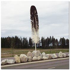 a large feather sculpture sitting on the side of a road next to rocks and trees