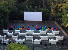 an outdoor movie theater with white chairs and large screen in the background, surrounded by trees