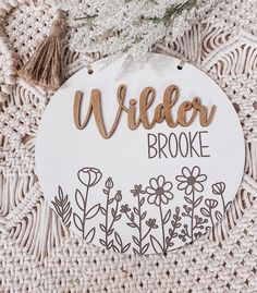 a wooden sign that says, wilder brooke surrounded by flowers and tassels on a crocheted surface