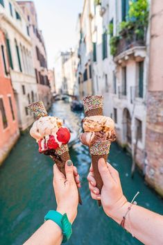 two people holding up ice cream cones in front of a canal with buildings on both sides