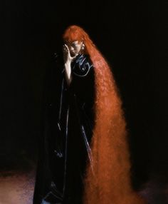 a woman with long red hair wearing a black cape and holding her hands to her face