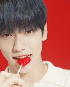 a young man with black hair holding a red lollipop in front of his mouth