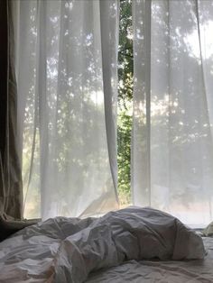 an unmade bed in front of a window with sheer curtains on the windowsill