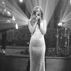 a woman in a long white dress holding a microphone and singing on stage with lights behind her