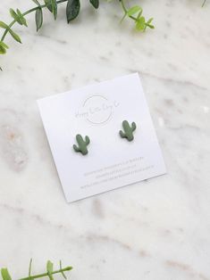 a pair of green cactus earrings sitting on top of a white marble table next to greenery