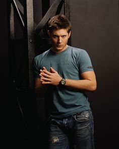 a young man standing in front of a black wall with his hands folded and looking at the camera