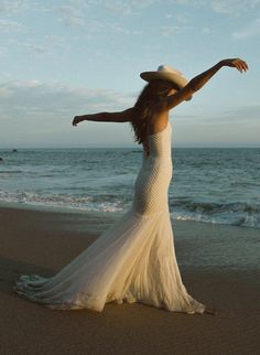 a woman in a white dress and hat standing on the beach with her arms outstretched