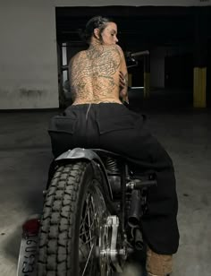 a tattooed man sitting on top of a motorcycle in a parking garage with his back turned to the camera