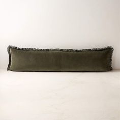 an olive green velvet pillow with fringe trim on the bottom and back, sitting against a white wall