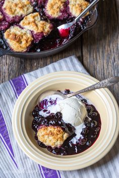 blueberry cobbler with ice cream in a bowl on a wooden table next to a cast iron skillet