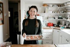 a woman standing in a kitchen holding a tray with food on it and smiling at the camera