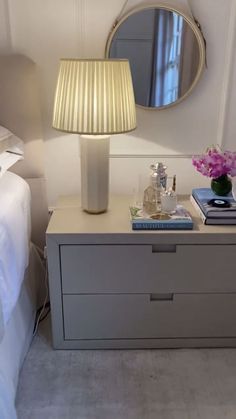 a nightstand with a lamp and mirror on top of it next to a white bed