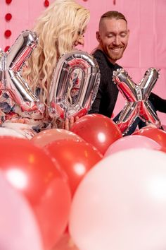 A valentines day backdrop and theme with a man and a woman behind pink and red balloons with silver letter balloons that say XOXO there's a pink back drop with red hearts on it. A valentines day backdrop Valentines Backdrop, Balloons Valentines Day, Valentine Backdrop, Backdrop Ideas, Photo Shoot