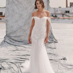 a woman standing in front of a backdrop wearing a wedding dress with an off the shoulder design